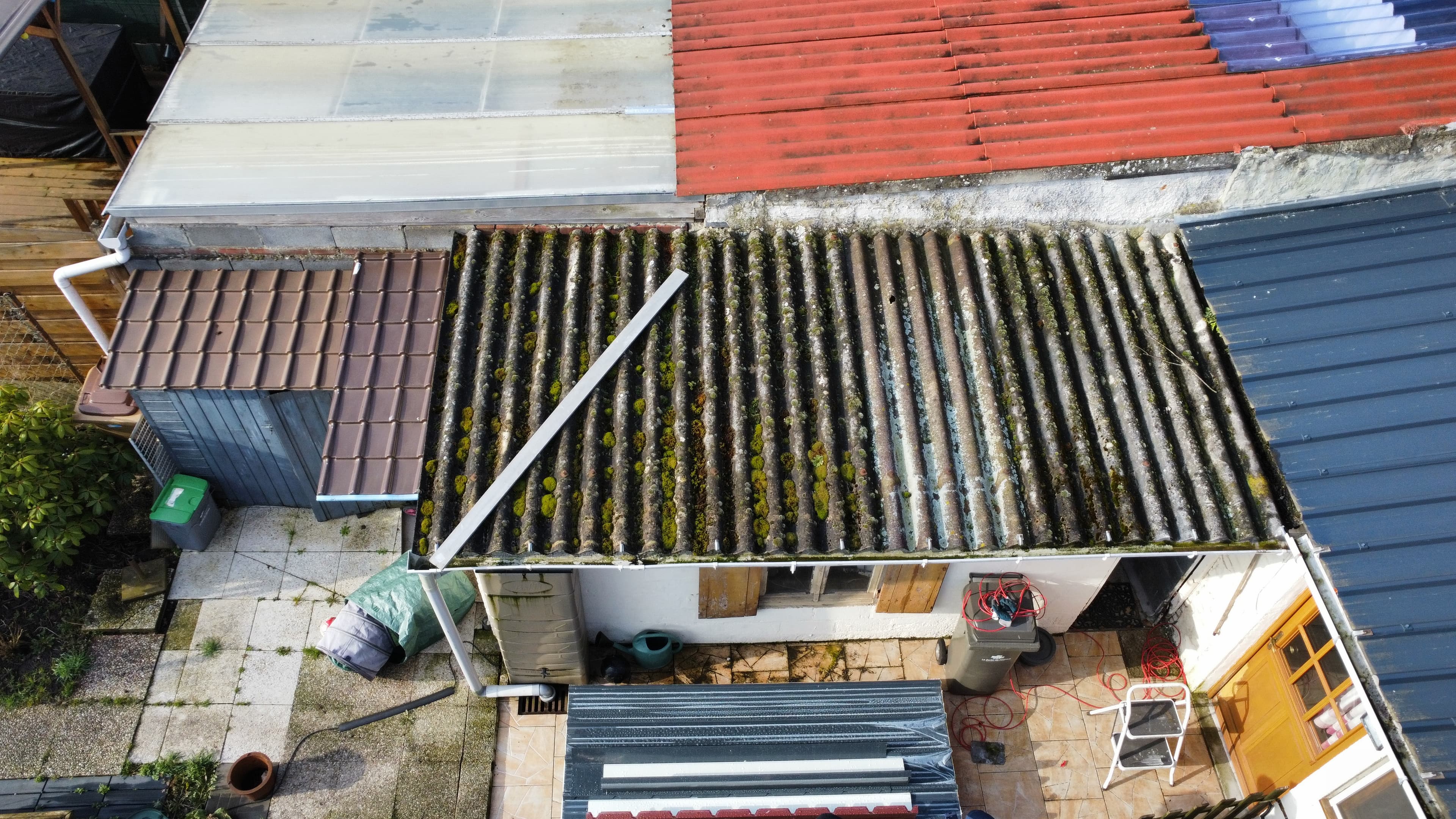 Vue plongeante sur divers toits, dont un toit ondulé gris parsemé de mousse verte.