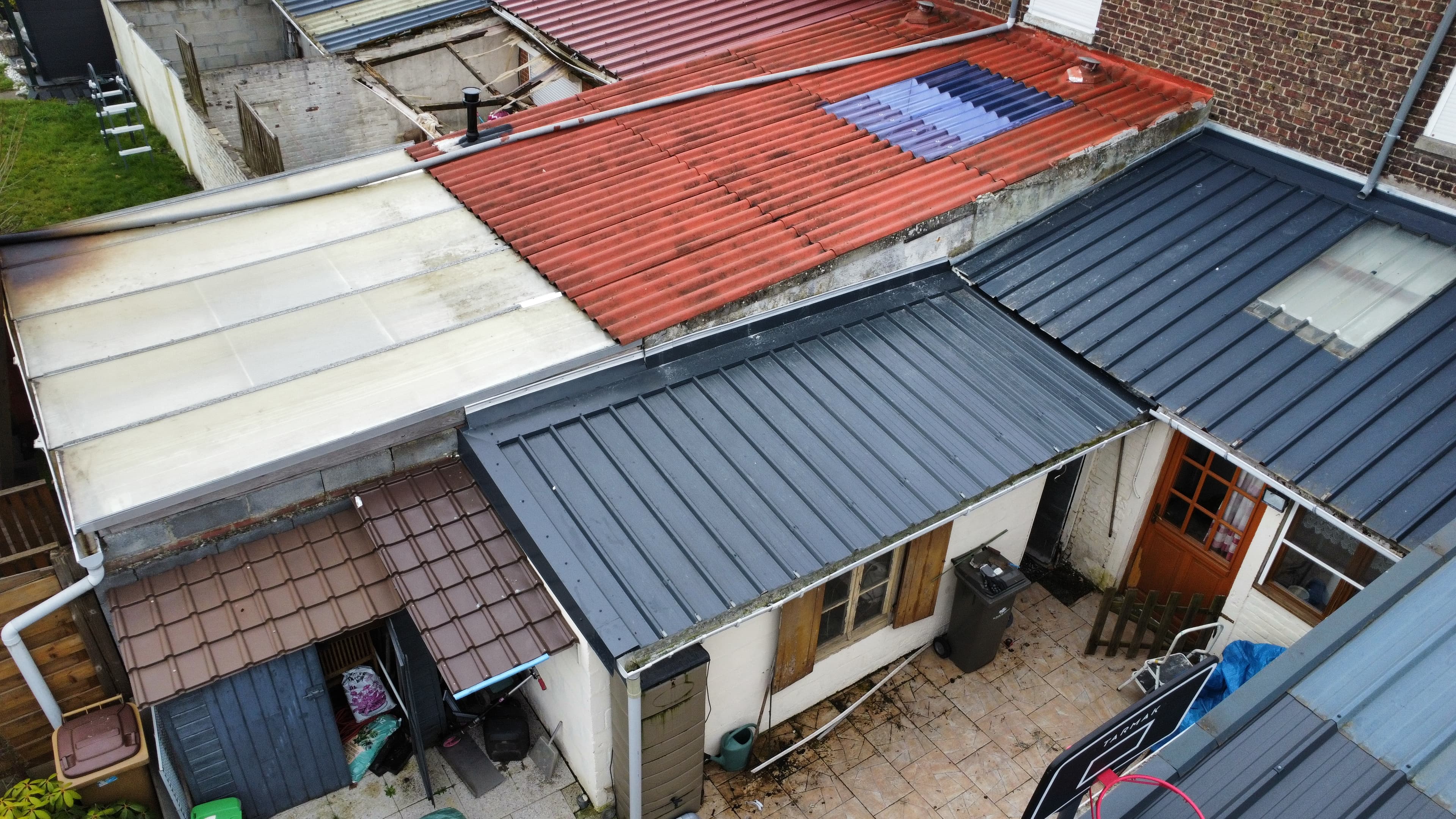 Toitures disparates en métal rouge, gris et plastique translucide surplombant une petite cour pavée.